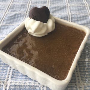 Square white ramekin filled with dark brown custard. The top is shiny from the caramelied sugar. There is a dollop of whipped cream and a chocolate heart in one corner. The ramekin sits on a blue and white plaid tablecloth.